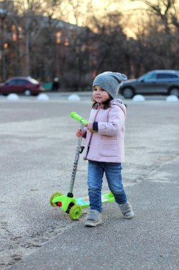 Şapkalı sevimli kız, palto ve kot pantolonla üç tekerlekli scooter'a biniyor. Akşam bahar şehir gün batımı.