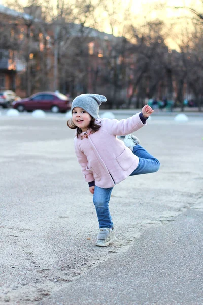 Schöne kleine Mädchen mit Hut, Mantel und Jeans springt, fliegt und hat Spaß auf dem Hintergrund des Stadtbildes im Licht des Sonnenuntergangs. — Stockfoto