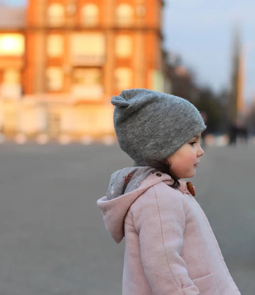 Avond portret van een mooi klein meisje in een hoed en vacht op de achtergrond van het stedelijk landschap in Sunset Light. — Stockfoto
