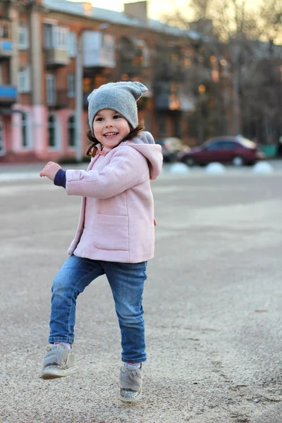 Mooi klein meisje in een hoed, jas en jeans springt, vliegt en heeft plezier op de achtergrond van het stadsbeeld in het licht van de zonsondergang. — Stockfoto