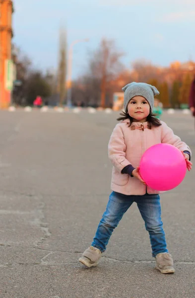 Klein wit meisje spelen met een bal op de straat in de avond. Portret op de achtergrond van de zonsondergang stadsgezicht — Stockfoto