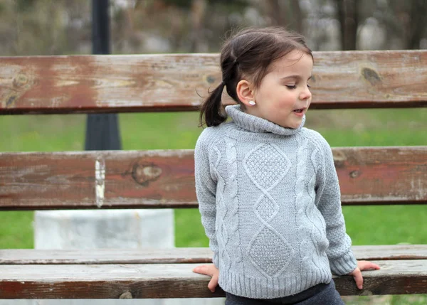 Piękna dziewczynka z ponytails i sweter siedzi na ławce w parku i uśmiechnięty — Zdjęcie stockowe