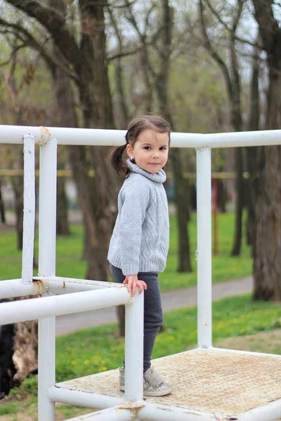 Kleines fröhliches brünettes Mädchen im Pullover und mit Frack steht auf einer Metallkonstruktion auf einer Baustelle — Stockfoto