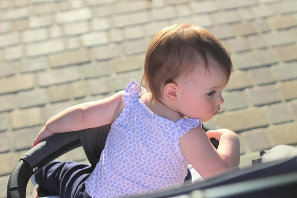 Pequeno, lindo, sorrindo, bebê ruiva bonito em uma camisa sem mangas em um carrinho fora de portas cai as mãos para baixo e olhando para trás — Fotografia de Stock