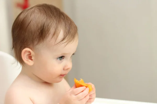 Das niedliche blonde Kind sitzt in einem Kinderstuhl in der Küche und isst einen saftigen Pfirsich. kleines Mädchen mit einem stechenden Blick — Stockfoto