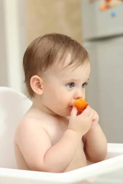 Das niedliche blonde Kind sitzt in einem Kinderstuhl in der Küche und isst einen saftigen Pfirsich. kleines Mädchen mit einem stechenden Blick — Stockfoto