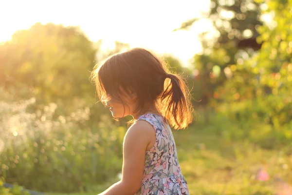 Silhouette of a little girl with a ponytail in a flower dress outdoors in the rays of sunny sunset — Stock Photo, Image