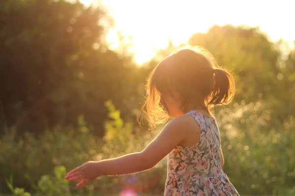 Silueta de una niña pequeña con una cola de caballo en un vestido de flores al aire libre en los rayos de sol puesta de sol — Foto de Stock