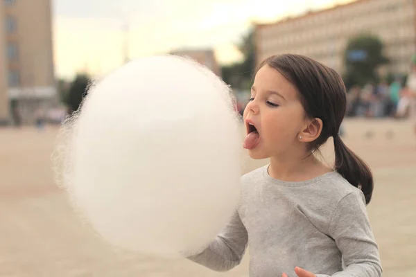 Little girl with pigtails tries to bite a big ball of cotton candy at a city celebration — Stock Photo, Image