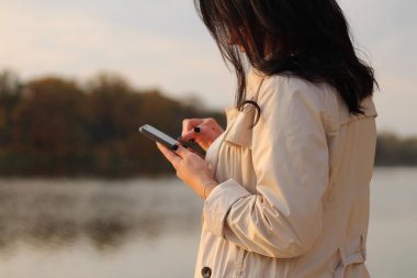 Beyaz siyah saçlı kız cep telefonuna bakar ve bir sonbahar gölünün arka planında telefon ekranına parmağını geçirir.