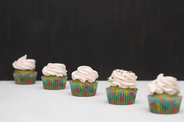 Cupcakes verdes com chapéu de creme em papéis multicores em uma mesa branca em um fundo escuro. Foco no bolo, profundidade do efeito de campo — Fotografia de Stock