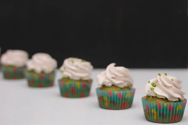 Pastelitos verdes con sombrero crema en papeles multicolores sobre una mesa blanca sobre un fondo oscuro. Enfoque en la torta, profundidad del efecto campo — Foto de Stock