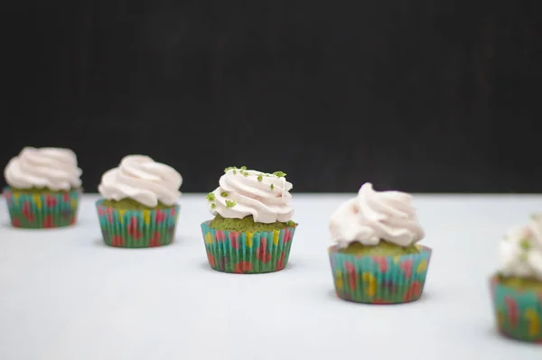 Pastelitos verdes con sombrero crema en papeles multicolores sobre una mesa blanca sobre un fondo oscuro. Enfoque en la torta, profundidad del efecto campo — Foto de Stock