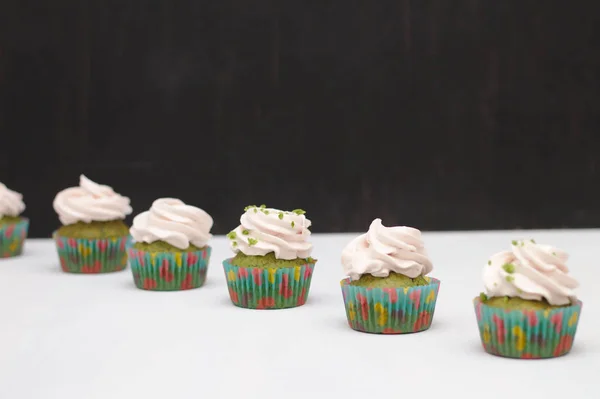 Green cupcakes with cream hat in multi-colored papers on a white table on a dark background. Focus on cake, depth of field effect — Stock Photo, Image