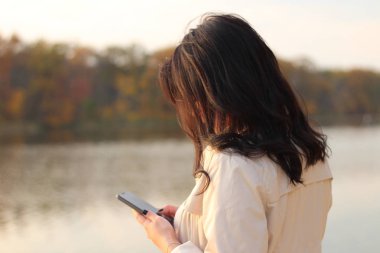 Beyaz siyah saçlı kız cep telefonuna bakar ve bir sonbahar gölünün arka planında telefon ekranına parmağını geçirir.