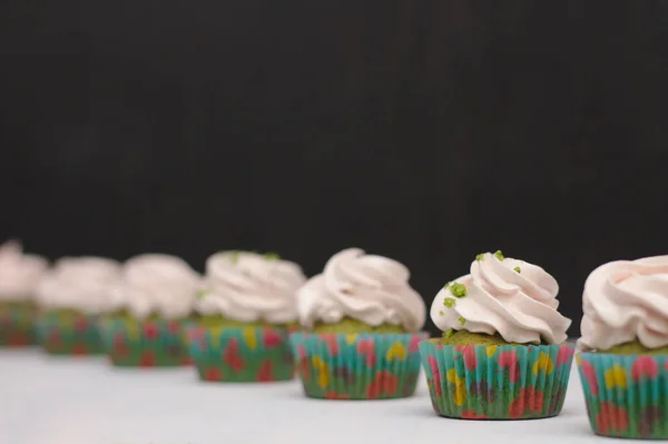 Cupcakes verdes com chapéu de creme em papéis multicores em uma mesa branca em um fundo escuro. Foco no bolo, profundidade do efeito de campo — Fotografia de Stock