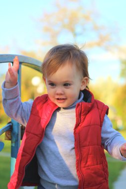 Kırmızı yelekli beyaz sarışın çocuk nehrin üzerindeki köprünün korkuluklarında asma kilit olduğunu düşünüyor. Sonbahar parkında yürü