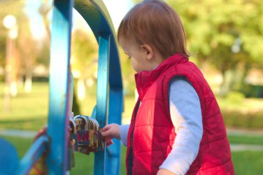 Kırmızı yelekli beyaz sarışın çocuk nehrin üzerindeki köprünün korkuluklarında asma kilit olduğunu düşünüyor. Sonbahar parkında yürü