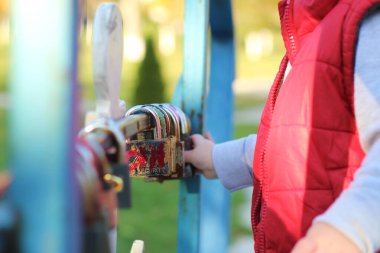 Kırmızı yelekli beyaz sarışın çocuk nehrin üzerindeki köprünün korkuluklarında asma kilit olduğunu düşünüyor. Sonbahar parkında yürü
