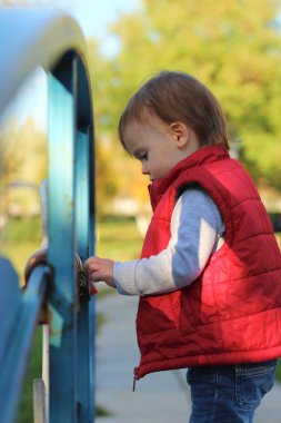 Kırmızı yelekli beyaz sarışın çocuk nehrin üzerindeki köprünün korkuluklarında asma kilit olduğunu düşünüyor. Sonbahar parkında yürü