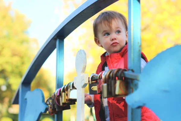 Niño rubio blanco en un chaleco rojo considerando candados en la barandilla del puente sobre el río. Caminar en el parque de otoño —  Fotos de Stock