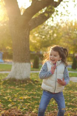 Neşeli bir çocuk bir kucak dolusu düşen sarı yaprakları saçıyor. Sonbahar parkında güneşli bir gün batımı.
