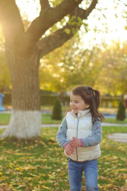 Neşeli bir çocuk bir kucak dolusu düşen sarı yaprakları saçıyor. Sonbahar parkında güneşli bir gün batımı.