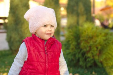 Pofuduk pembe şapkalı ve kırmızı yelekli küçük bir çocuk sonbahar parkında bir çam ağacının yanında duruyor. Güzel sonbahar açık havada güneşli bir gün.