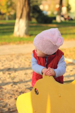 Pembe şapkalı ve kırmızı yelekli küçük bir çocuk salıncağın yanındaki sonbahar parkında duruyor. Güzel sonbahar açık havada güneşli bir gün.
