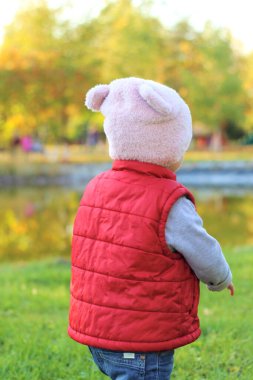 Pembe şapkalı ve kırmızı yelekli küçük bir çocuk arkasında duruyor. Çocuk kıyıda durur ve sonbahar gölüne bakar.