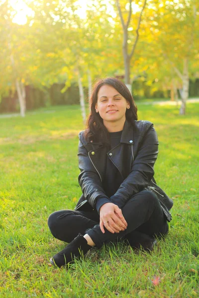 Uzun siyah saçlı beyaz kız, sonbahar sarı ağaçlarının ve yaprakları dökülmüş çimenlerin arka planında deri bir ceket içinde oturuyor.