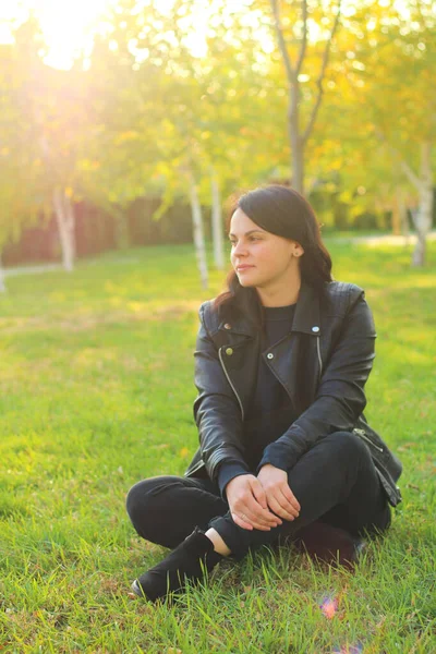 Chica blanca con el pelo largo y oscuro está sentado en una chaqueta de cuero sobre un fondo de árboles amarillos de otoño y hierba con hojas caídas —  Fotos de Stock