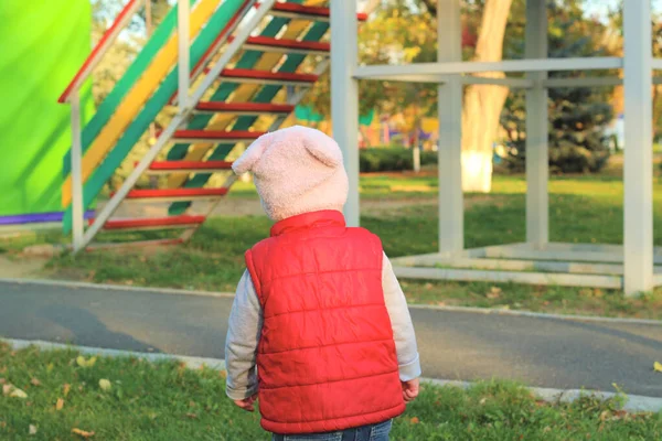 Ένα μικρό παιδί με ένα χνουδωτό ροζ καπέλο και κόκκινο γιλέκο τρέχει στο πάρκο του φθινοπώρου. Όμορφη ηλιόλουστη μέρα σε εξωτερικούς χώρους. — Φωτογραφία Αρχείου