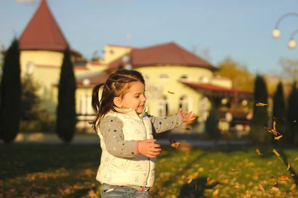 Un bambino allegro disperde una manciata di foglie gialle cadute. Tramonto soleggiato nel parco autunnale all'aperto — Foto Stock