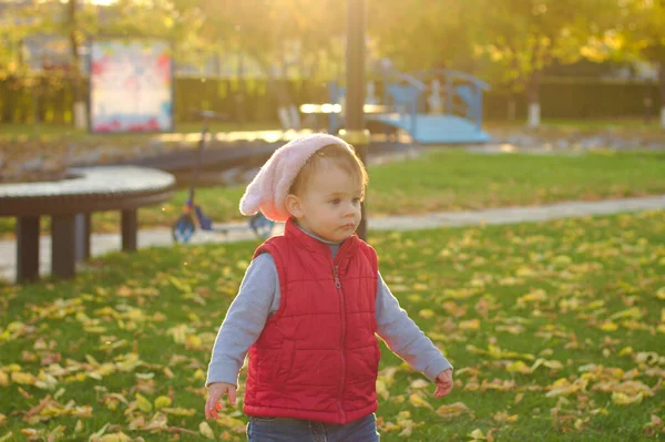 Een klein kind met een pluizige roze hoed en rood vest loopt in het herfstpark. Mooie zonnige herfstdag buiten. — Stockfoto