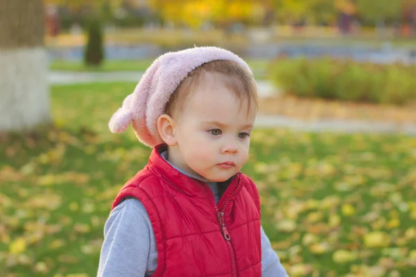Egy kis gyerek bolyhos rózsaszín kalapban és piros mellényben szaladgál az őszi parkban. Gyönyörű őszi napsütéses nap a szabadban. — Stock Fotó