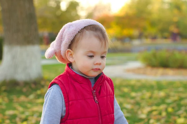 Un niño pequeño con un sombrero rosa esponjoso y un chaleco rojo está corriendo en el parque de otoño. Hermoso otoño día soleado al aire libre . — Foto de Stock