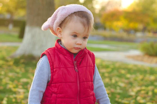 Un niño pequeño con un sombrero rosa esponjoso y un chaleco rojo está corriendo en el parque de otoño. Hermoso otoño día soleado al aire libre . — Foto de Stock