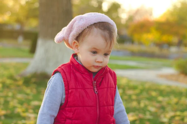 Un niño pequeño con un sombrero rosa esponjoso y un chaleco rojo está corriendo en el parque de otoño. Hermoso otoño día soleado al aire libre . — Foto de Stock