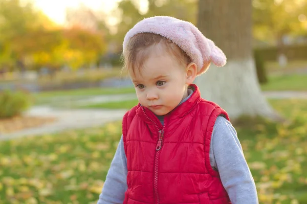 Un niño pequeño con un sombrero rosa esponjoso y un chaleco rojo está corriendo en el parque de otoño. Hermoso otoño día soleado al aire libre . — Foto de Stock