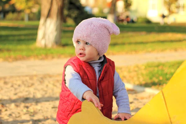 Een klein kind met een pluizige roze hoed en rood vest staat in het najaarspark vlakbij schommel. Mooie zonnige herfstdag buiten. — Stockfoto