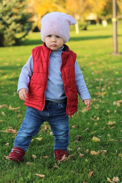 Een klein kind met een pluizige roze hoed en rood vest loopt in het herfstpark. Mooie zonnige herfstdag buiten. — Stockfoto