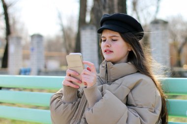 Şaşırmış bir kız bankta oturur ve telefonuna bakar.