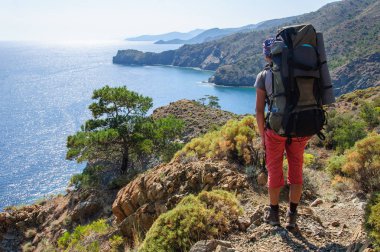 Sırt çantalı bir adam Akdeniz kıyısında duruyor ve uzaklığa bakıyor.