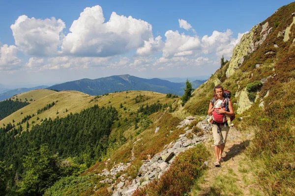 A girl with baby in a sling goes at the mountain range. Ukraine. Carpathian mountains — Stock Photo, Image