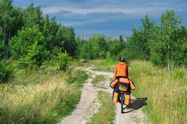A cyclist on sports bike rides with a large backpack along a dirt road through a birch grove — Stock Photo, Image