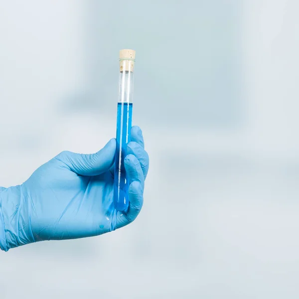 Mão em luvas segura um tubo de ensaio com líquido azul na frente de um fundo de laboratório — Fotografia de Stock