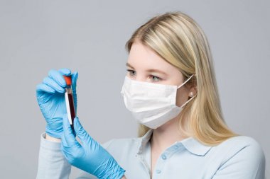 Tıbbi eldivenleri ve tıbbi maskesi olan genç bir kadın elinde bir kan sondası tutuyor.