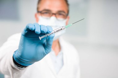 Elinde Covid-19 serumu / aşısı olan bir şırıngayla arka planda eldiven ve yüz maskesi olan bir doktor.