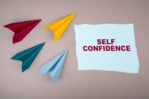 SELF CONFIDENCE (englanniksi) Teksti muistilehtiö, paperi lentokoneita ympäri, symboli saada tavoitteita — kuvapankkivalokuva